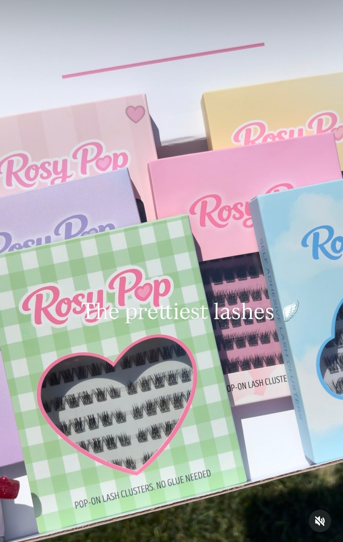 Several Rosy Pop pop-on lash cluster boxes arranged together, including pastel pink, yellow, lavender, green-and-white gingham, and light blue packages. A heart-shaped window on the front package reveals black lash clusters inside.