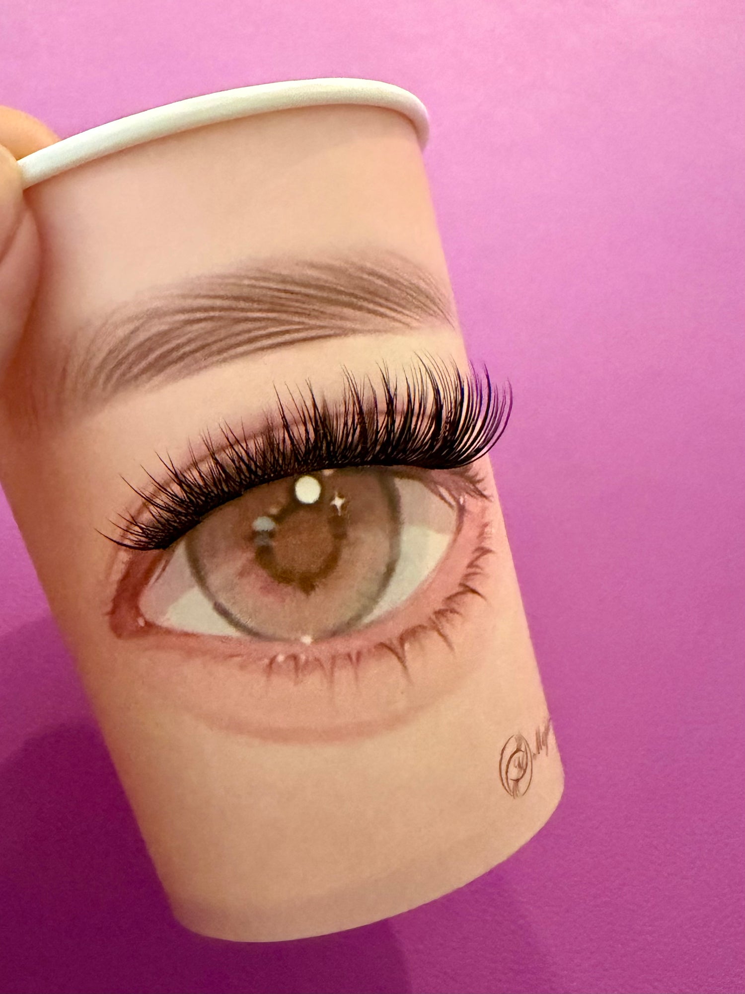 A close-up of a hand holding a paper cup printed with a realistic illustrated eye, eyebrow, and long false eyelashes, against a solid purple background.